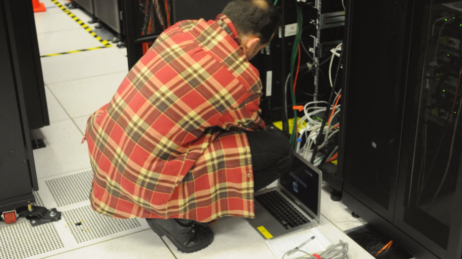 A technician works with a laptop beside network cables and server racks in a data center.