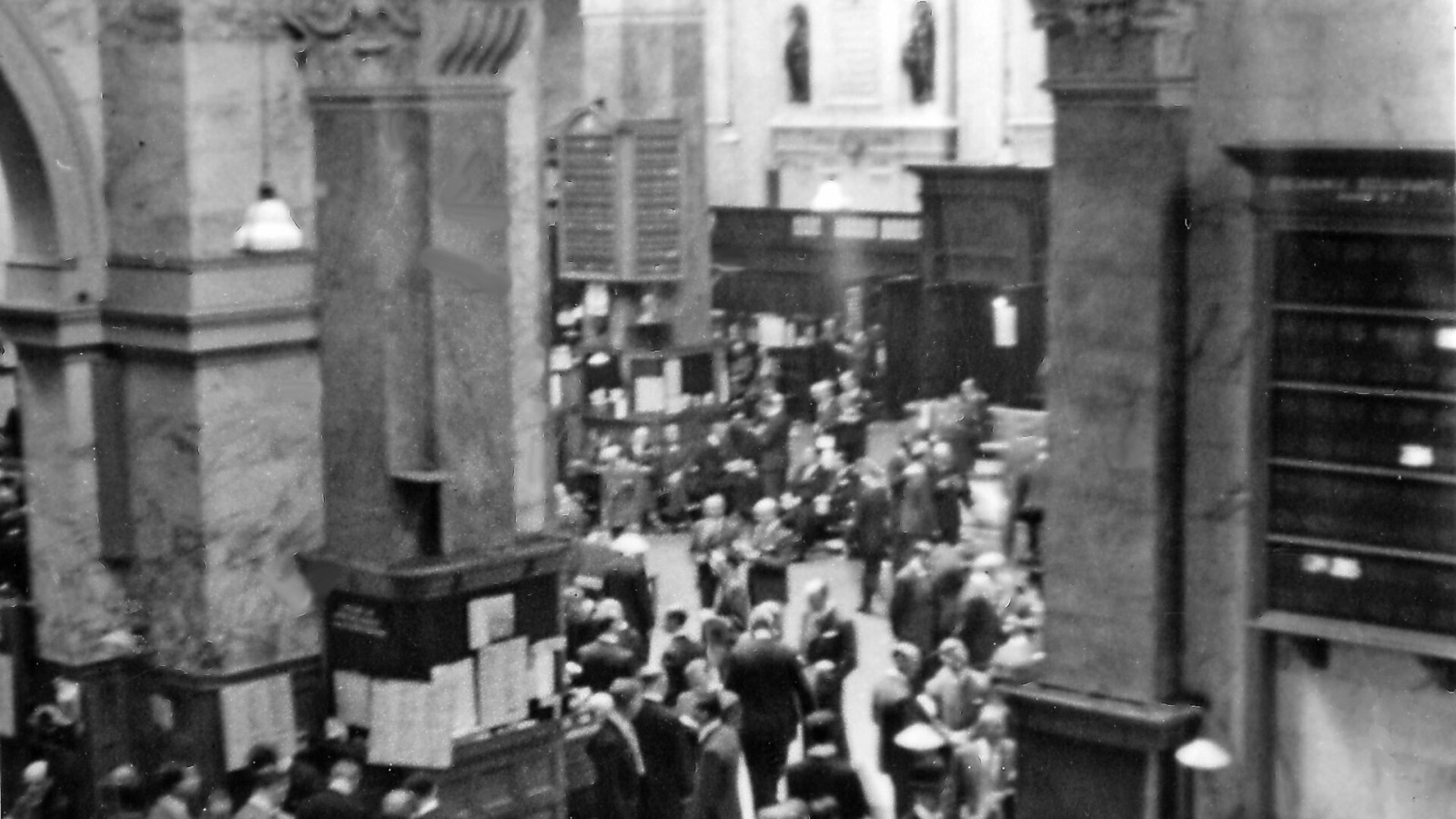 A historic black-and-white photograph of the London Stock Exchange trading floor filled with market participants