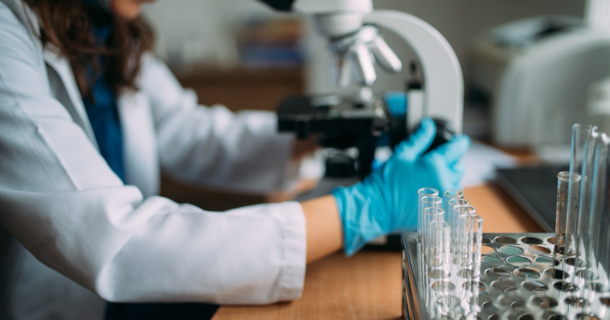 Une scientifique en blouse blanche et gants bleus travaille avec un microscope à côté de tubes à essai en verre.