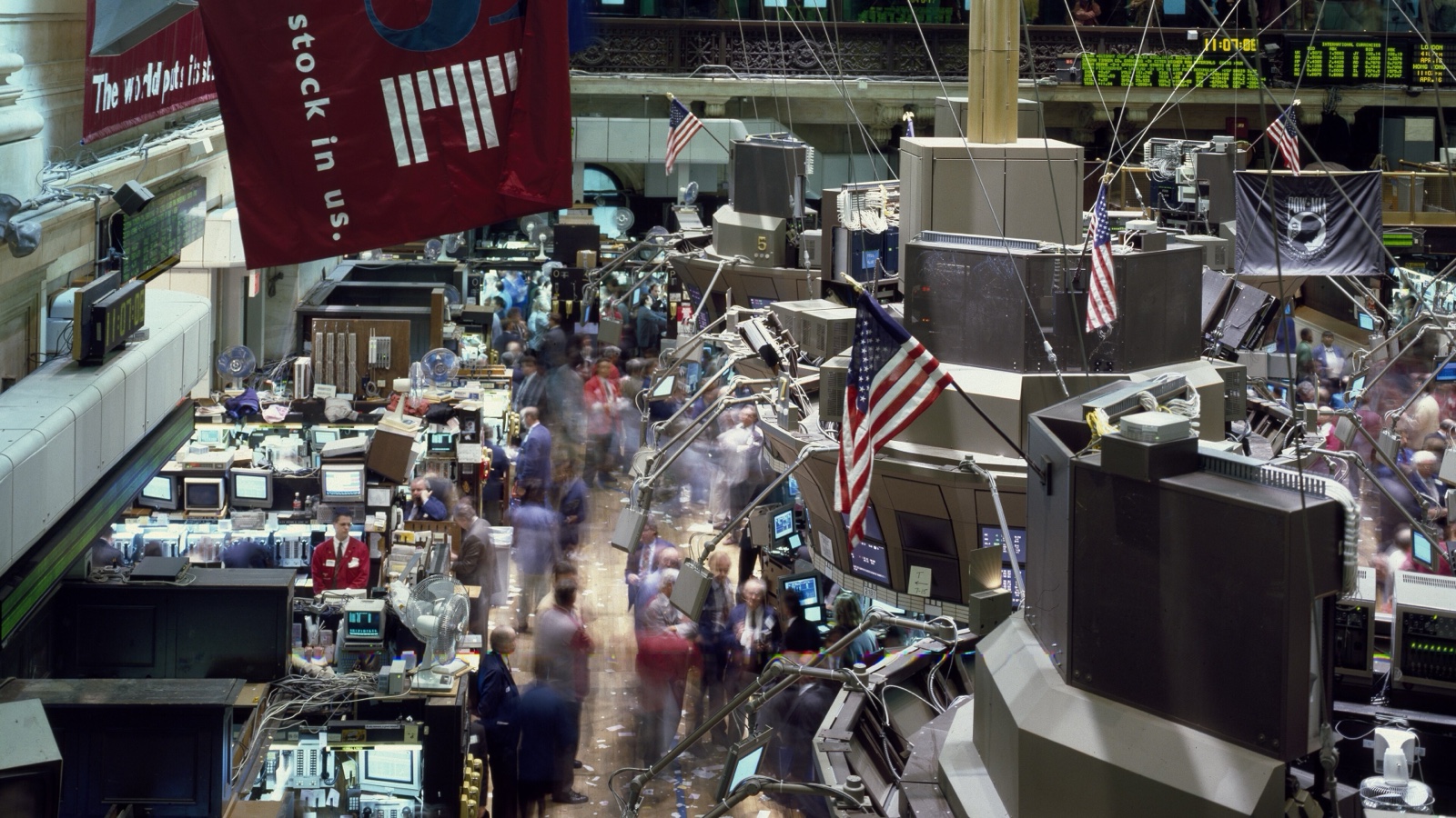 A busy New York Stock Exchange floor with moving traders, workstation posts, quote boards and American flags