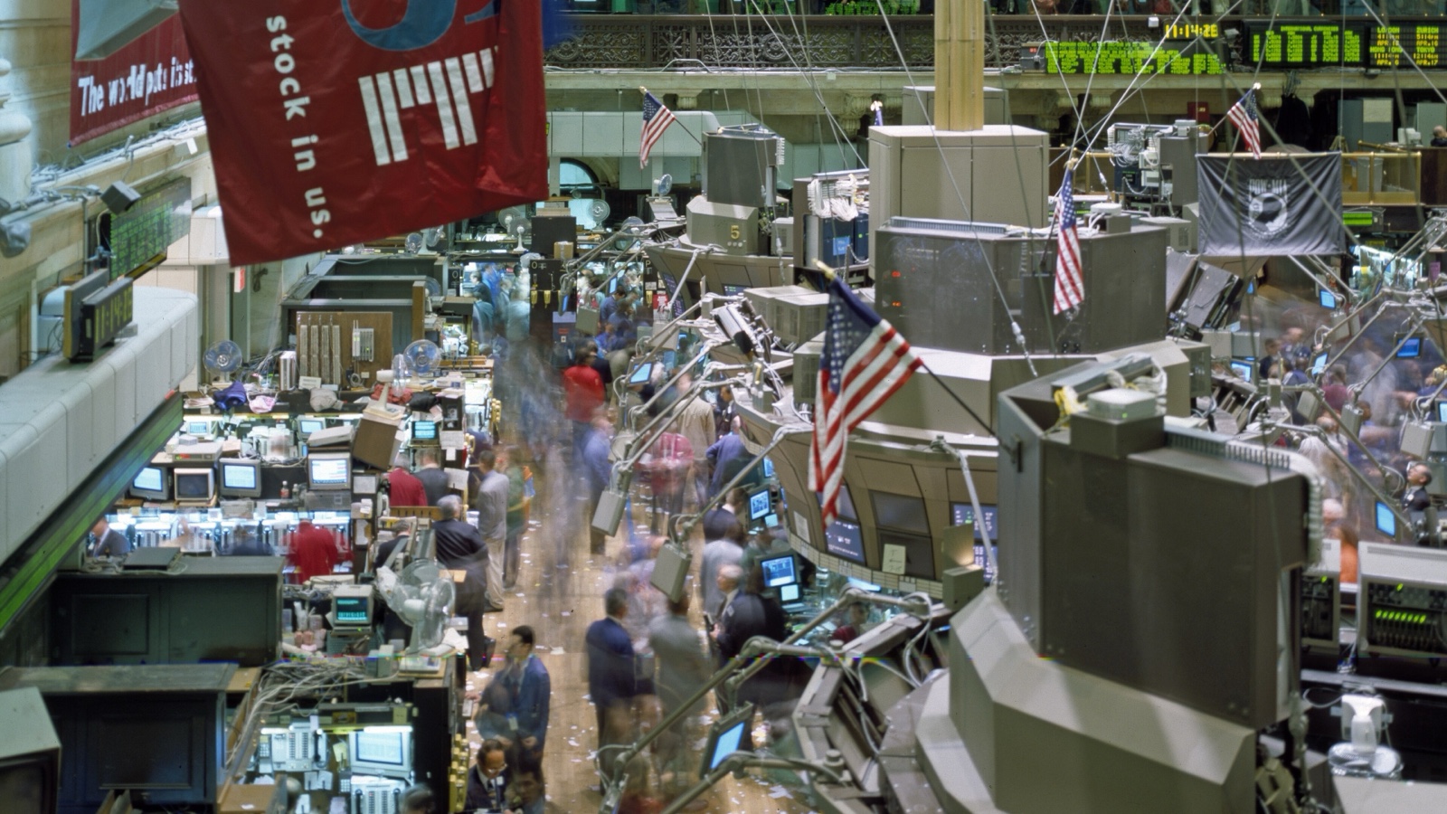A busy New York Stock Exchange floor with moving traders, workstation posts, quote boards, American flags and a large red market banner