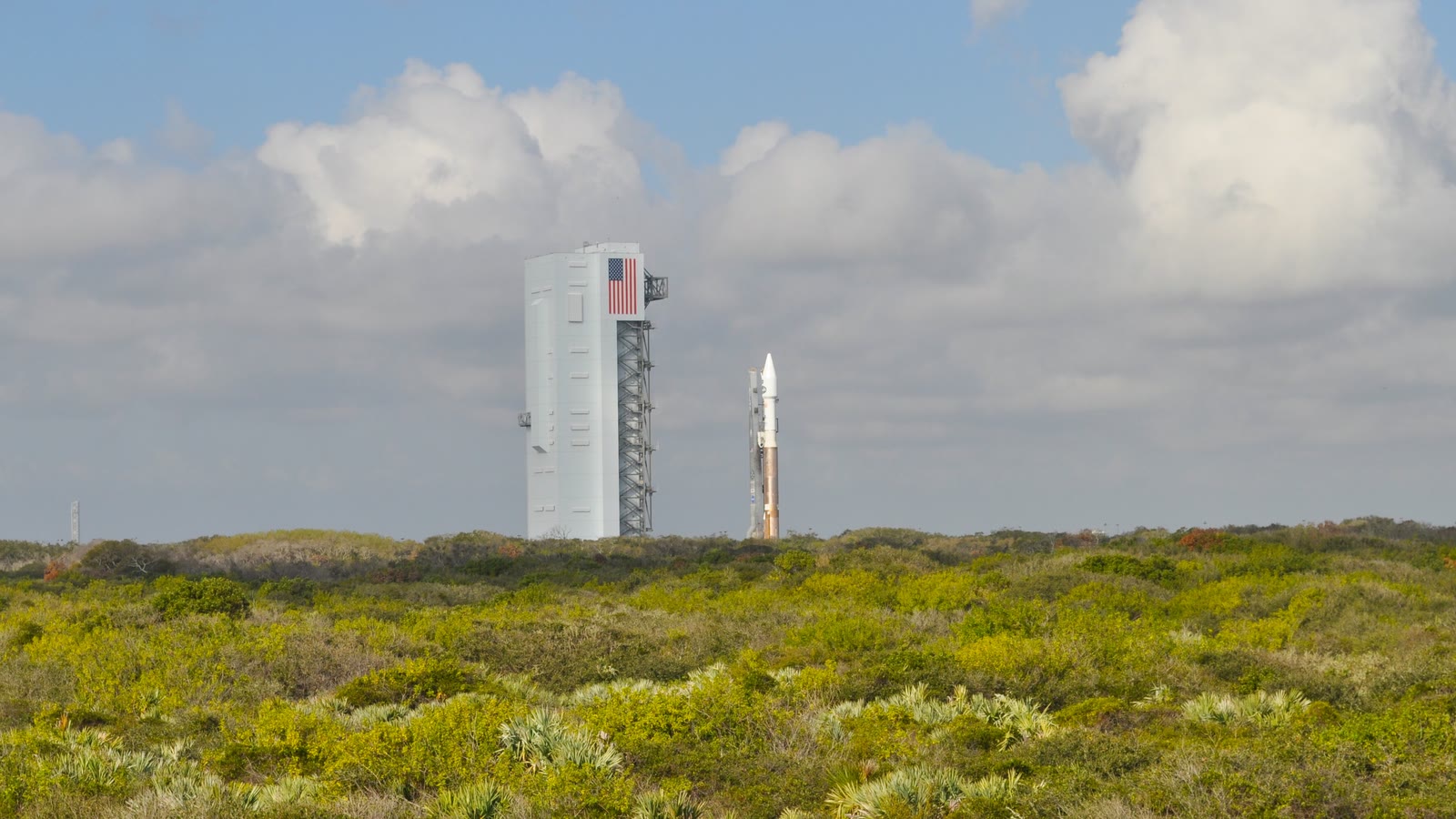 A communications-satellite rocket stands beside a launch tower at Cape Canaveral under a cloudy sky