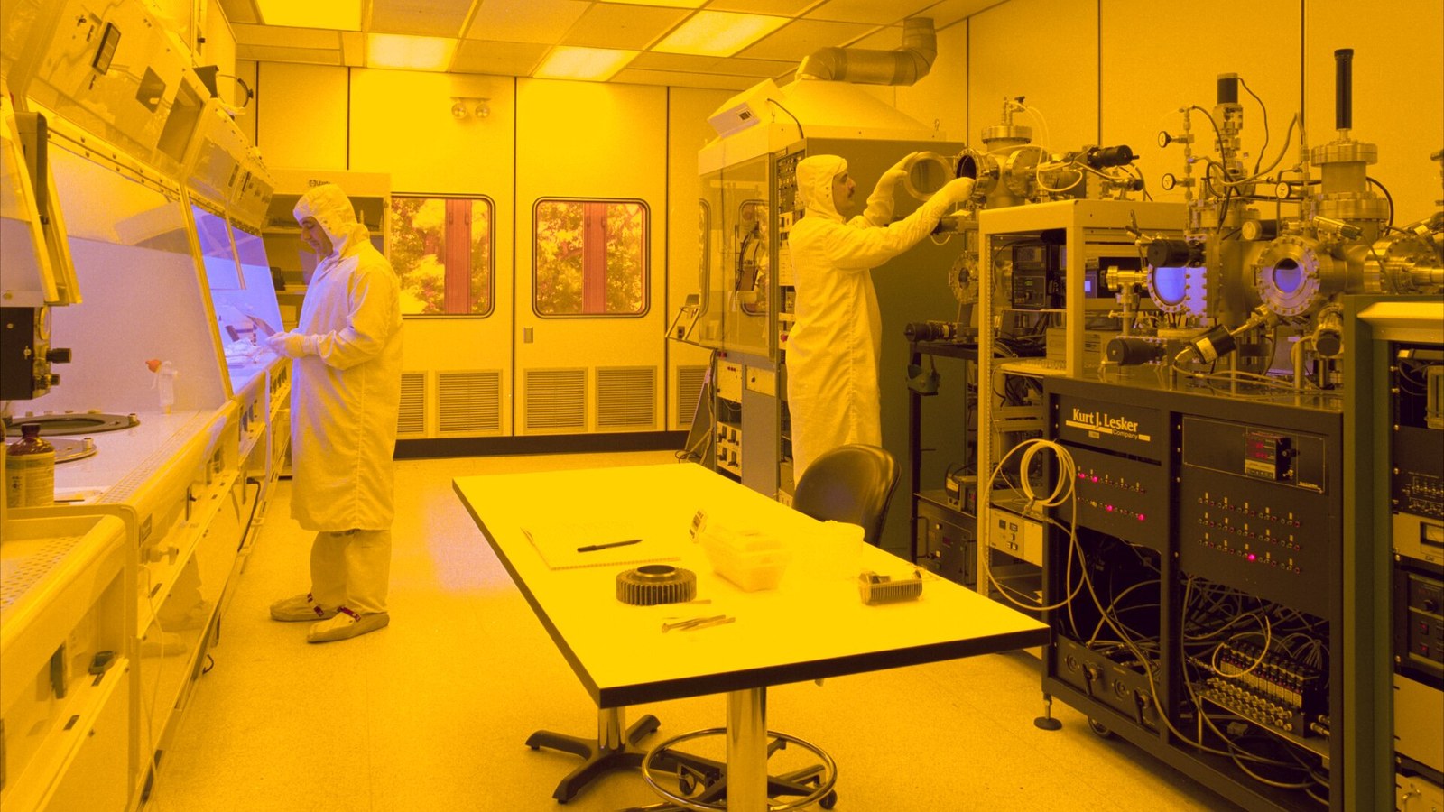 Two suited technicians work among semiconductor fabrication and vacuum equipment in a yellow-lit cleanroom