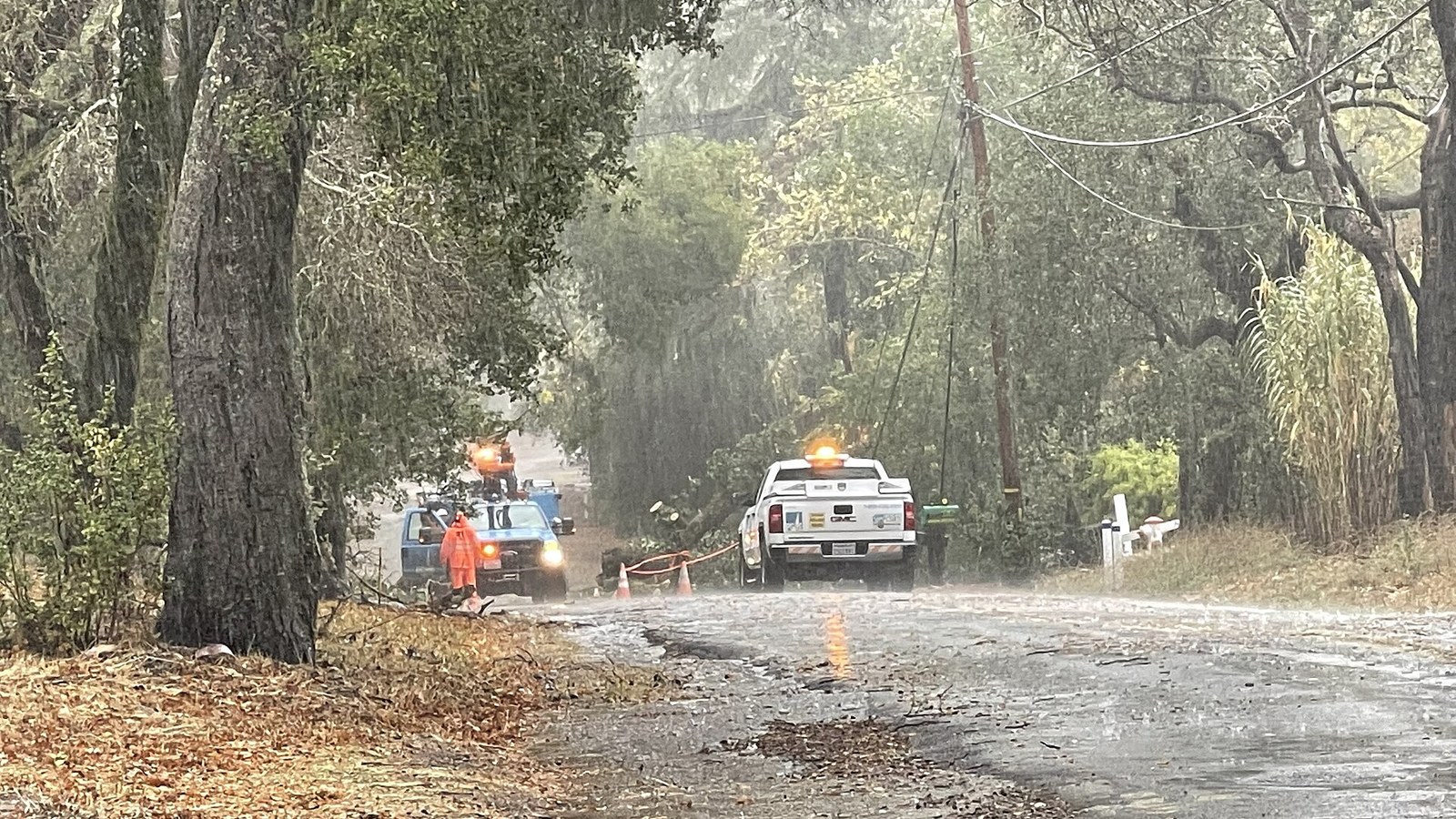 Two PG&E utility trucks and a high-visibility worker respond to downed lines on a rain-soaked California road