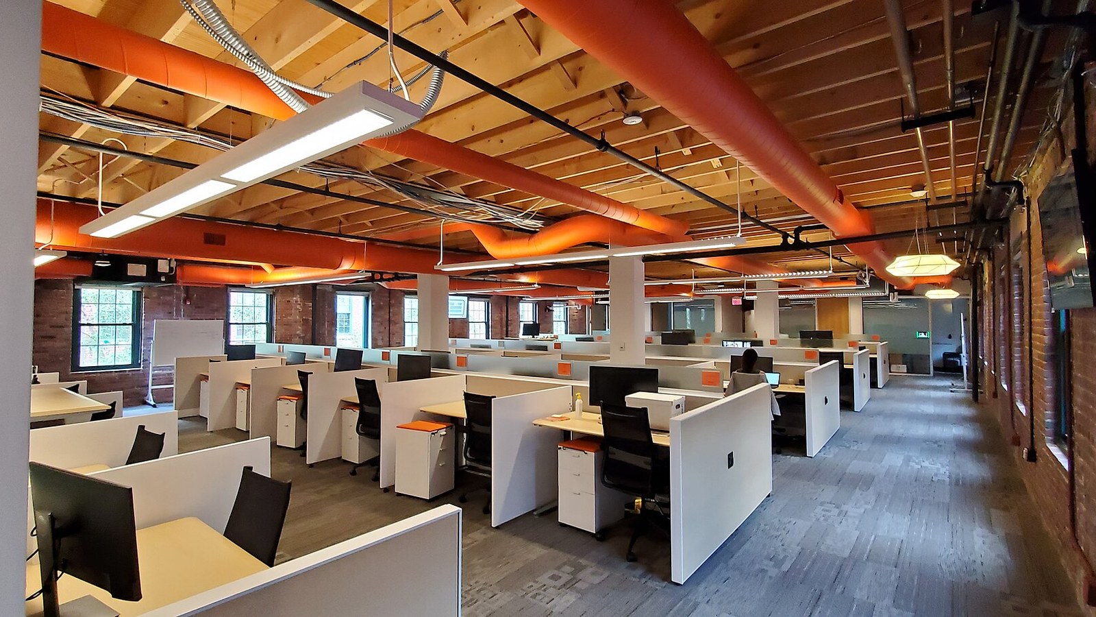 Des rangées de bureaux au siège de HubSpot à Cambridge, avec des poutres en bois et des conduits de ventilation orange