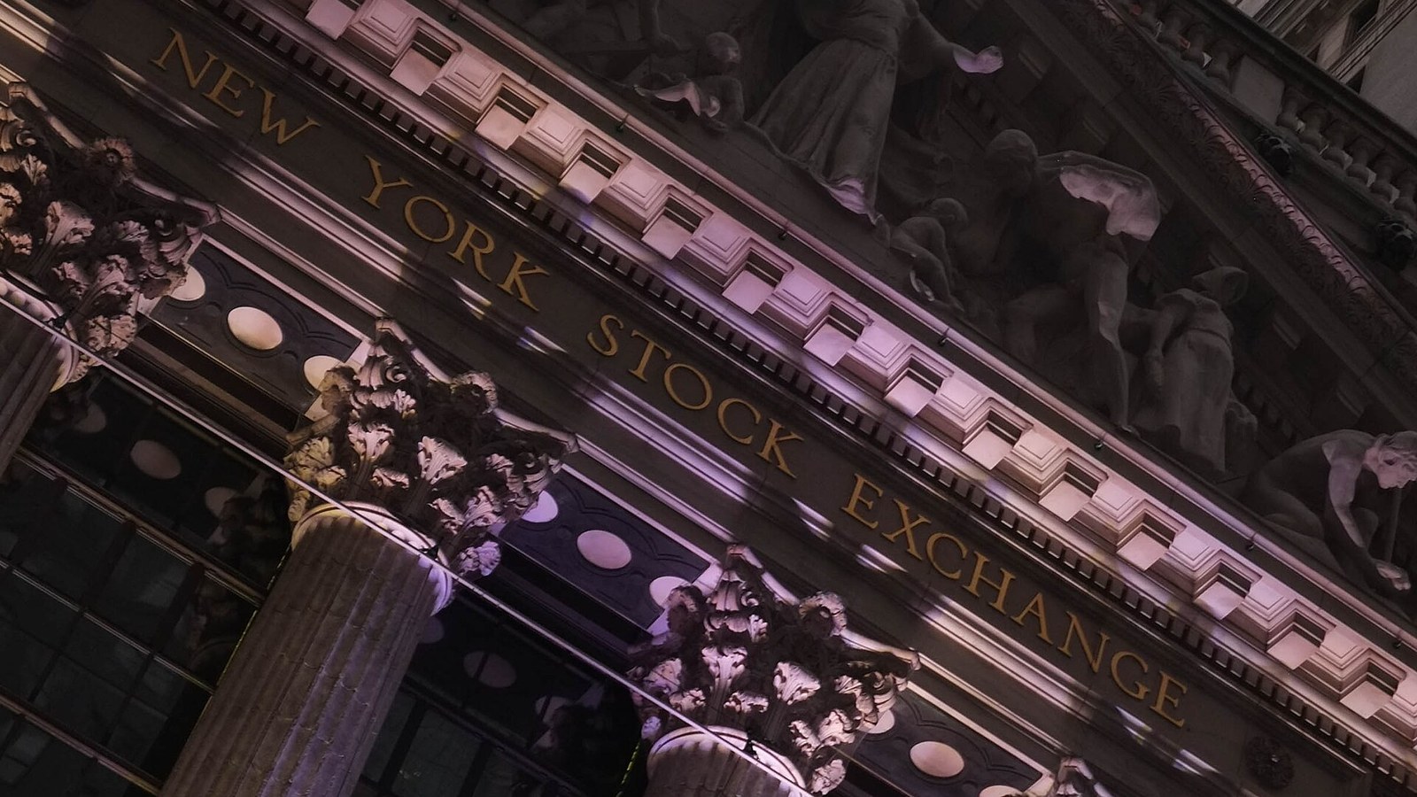 Angled close-up of the New York Stock Exchange façade, with gold lettering, ornate stone columns and sculpted figures in purple-toned light.