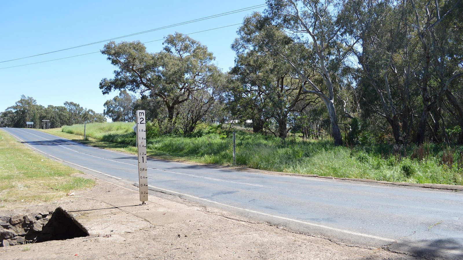 A white flood-depth marker post beside a rural road near Billabong Creek, Parkes, New South Wales