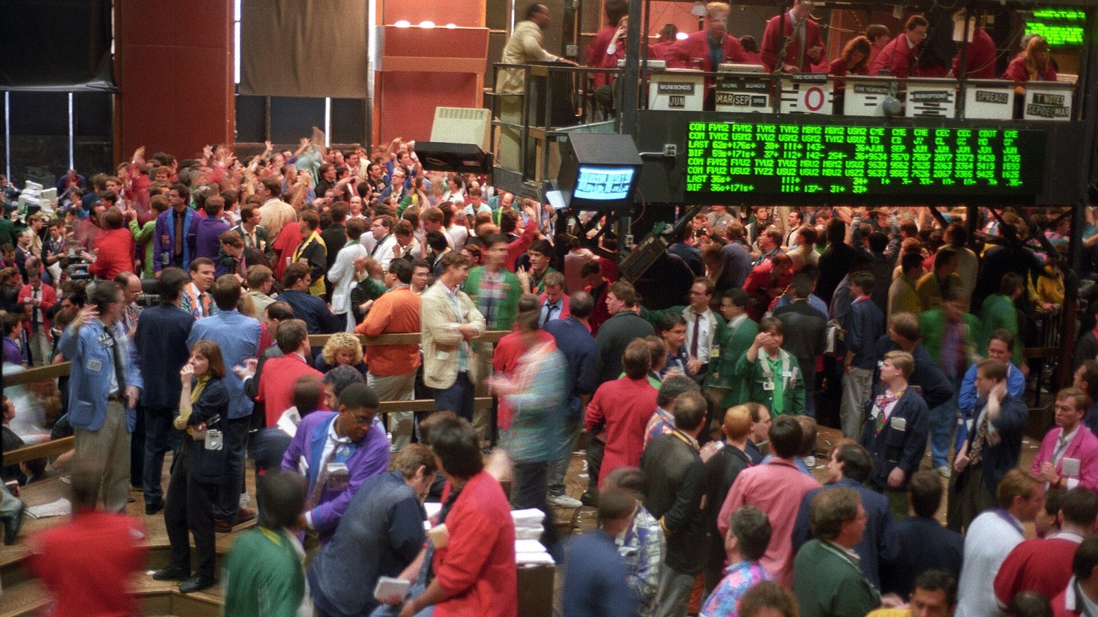 Traders crowd a financial exchange floor beneath large electronic market displays