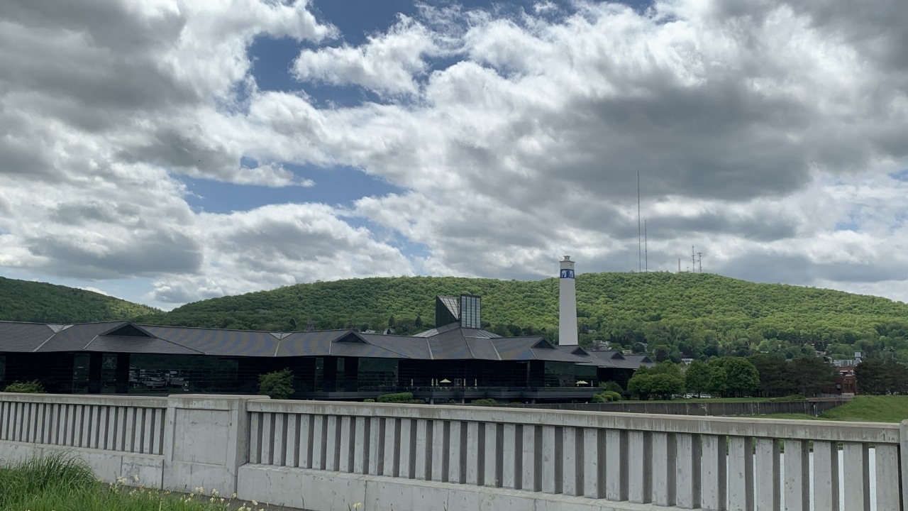 Photo d’archives de mai 2021 : bureaux de Corning et Little Joe Tower blanche, collines boisées, ciel nuageux et rambarde de béton au premier plan.