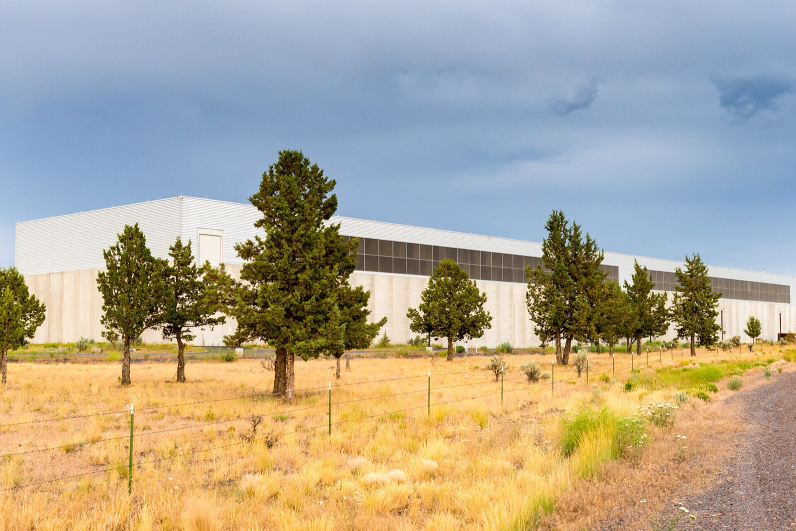 Un long centre de données blanc de Meta, derrière une rangée de genévriers et des herbes sèches, sous un ciel orageux à Prineville, en Oregon