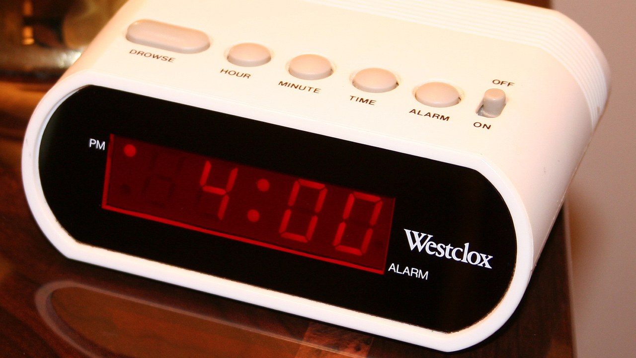 Close-up of a white digital alarm clock with a red 4:00 display on a reflective brown tabletop.