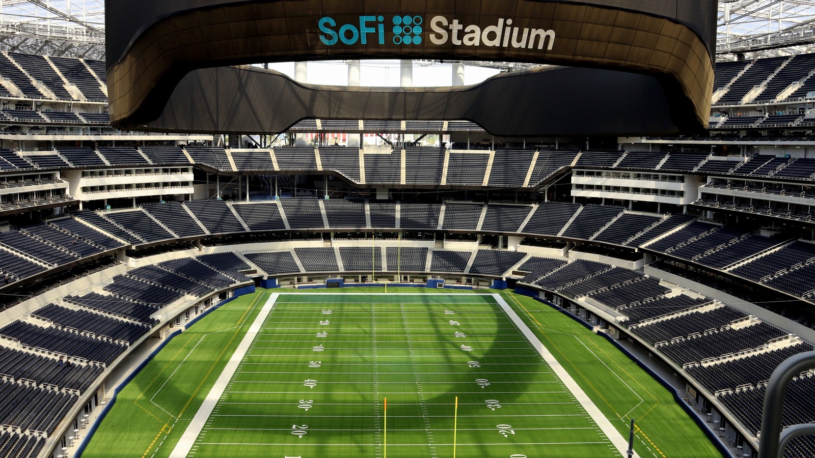 Archival March 2021 interior photograph of SoFi Stadium: an empty football field, blue seats and the SoFi Stadium name on the overhead display.