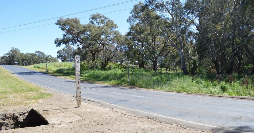 A white flood-depth marker post beside a rural road near Billabong Creek, Parkes, New South Wales