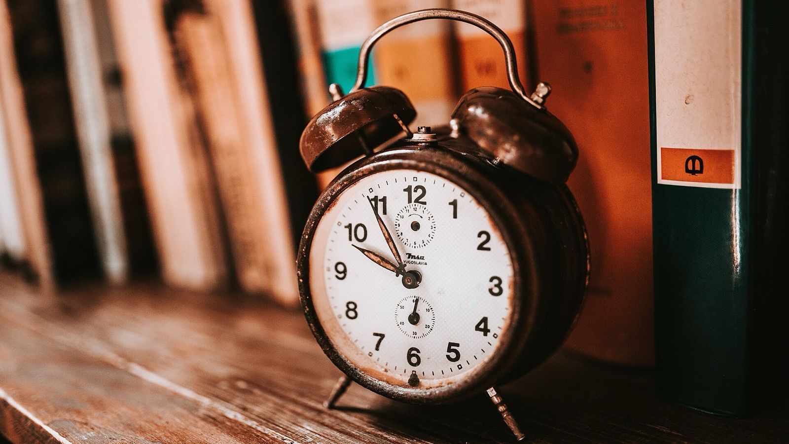 An old twin-bell alarm clock standing on a wooden bookshelf in front of books.